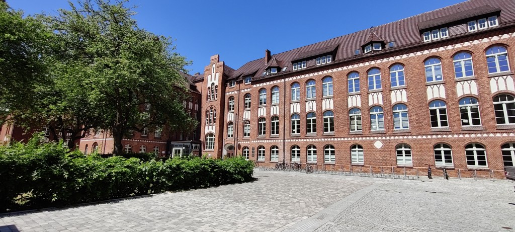 Kuvassa pitkä, vanha, nelikerroksinen tiilitalo, jonka edessä kivetty piha ja pensasaita sekä puita.