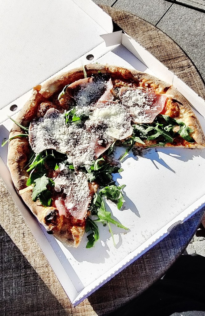 Kuvassa 3/4 pitsaa avonaisessa pitsalaatikossa. Pitsassa on parmankinkkua ja rucolaa, ja sen päälle on ripoteltu parmesaania.