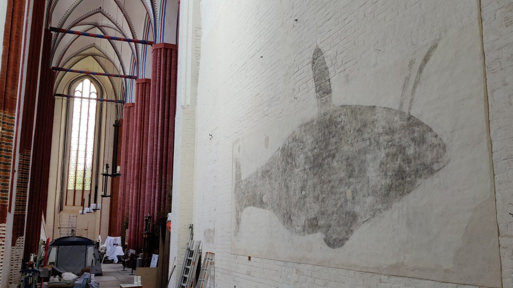 Kuva kirkon valkoisesta seinästä, johon on maalattu tummalla valtavan kokoinen kalamainen hahmo, jolla on tuntosarvet. Taustalla näkyy korkea kirkkosali ja remontointitarvikkeita.