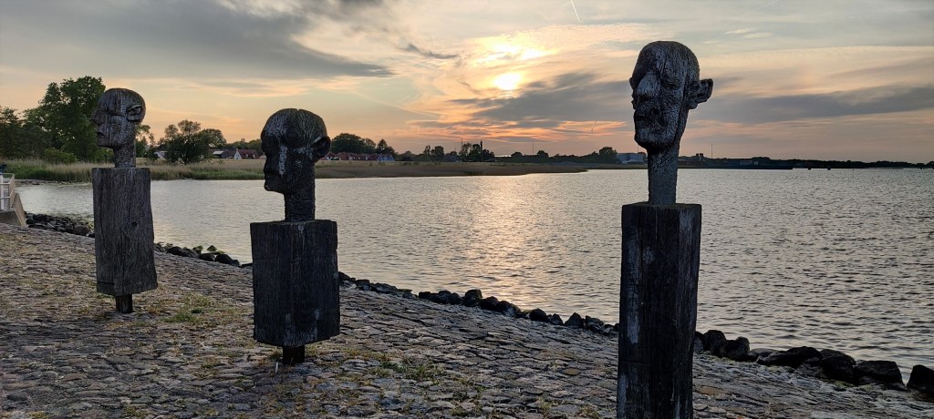 Kuvassa kolme ihmisen päätä esittävää patsasta, jotka seisovat silmät kiinni rivissä meren rannalla. Taustalla aurinko laskee pilvien taakse.