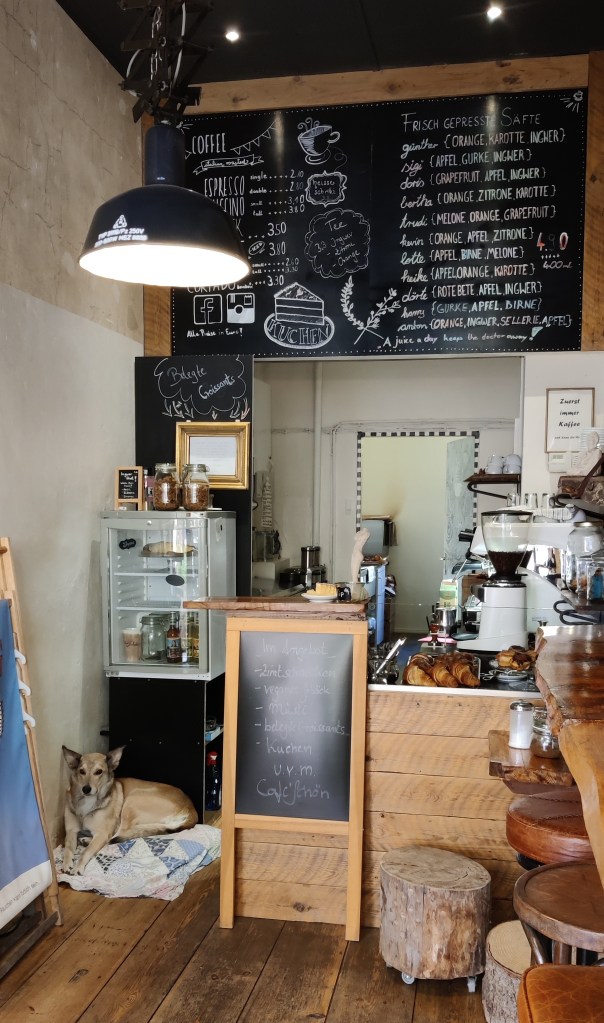 Kuva kahvilan kapeasta sisätilasta. Ylhäällä on iso liitutaulu, jossa esitellään tarjolla olevat tuotteet. Etualalla pieni tiski, jolla useampi croissant. Etuoikealla puisia jakkaroita ja seinän vieressä oleva kapea pöytä. Lattialla pötköttelee vaaleanruskea, pystykorvainen koira.