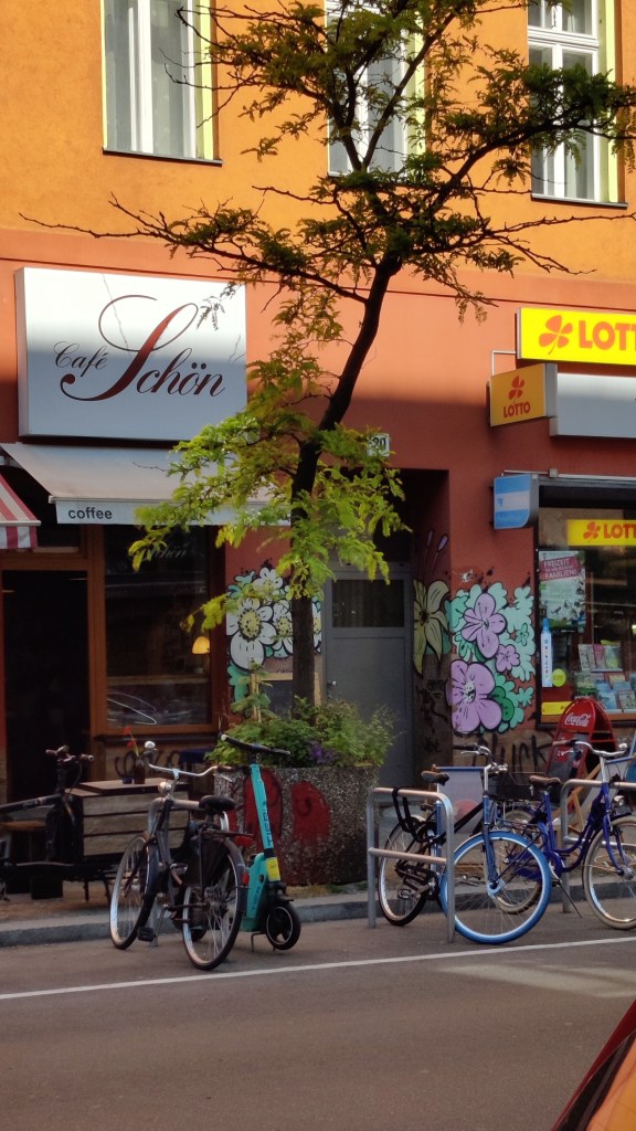 Kadulta otettu kuva Café Schönin julkisivusta. Edessä on puu ja useita polkupyöriä. Seinissä on sarjakuvamaisia kukkamaalauksia.