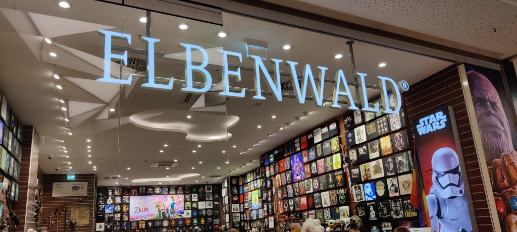 Kuva Elbenwald-kaupan näyteikkunasta. Oikeassa reunassa näkyy iso kuva Thanoksesta, sen vieressä Star Warsin trooper. Kaupan seinät ovat täynnä nörttikuvia painetuista t-paidoista. Kaukana takaseinällä näkyy Legend of Zelda -mainos.