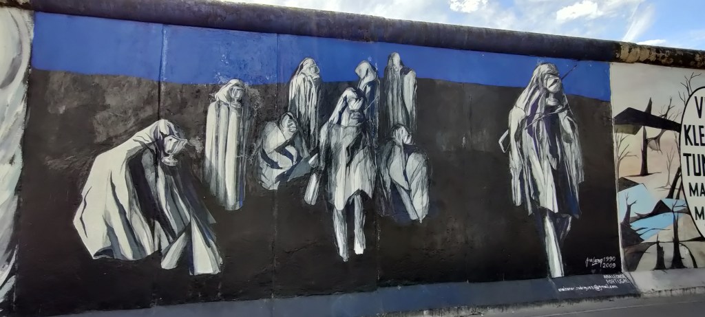 Kuvassa useita, suojaviittoihin ja happinaamareihin pukeutuneita ihmishahmoja. Hahmot ovat valkoisia, tausta musta, sininen taivas taustalla.