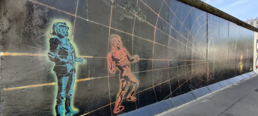 Vinosti otettu kuva taideteoksesta, jonka keskiössä näkyy epäselvä ihmishahmo. Kuvan etualalla on kaksi ihmishahmoa, toinen on pukeutunut futuristiseen haarniskaan ja toinen on alasti. Kumpikin on tehty ja varjostettu sarjakuvatyylillä.