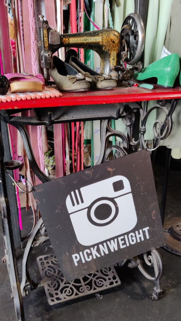 Kuva vanhanaikaisesta, poljettavasta Singerin ompelukoneesta, jonka päälle on aseteltu naisten kenkiä. Takana on kirjava vaaterekki ja etualalla roikkuu kyltti, jossa Instagramin kameralogo sekä teksti PICKNWEIGHT.