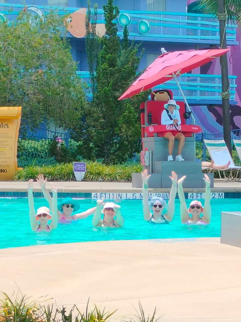 Kuva viidestä henkilöstä uima-altaassa. Kaikilla on päähineet ja aurinkolasit, ja jokainen nostaa molemmat kätensä ilmaan eri tavoin. Taustalla altaan reunalla istu punaisen päivänvarjon alla hengenpelastaja.