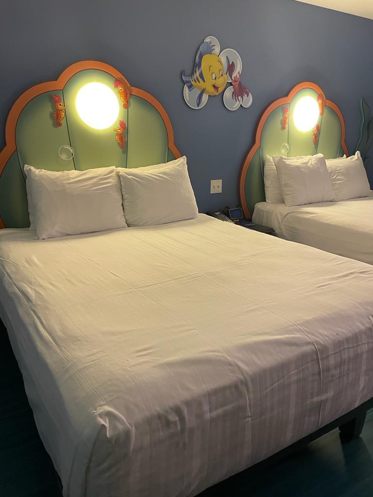 Kuvassa kaksi leveää parisänkyä, joissa kummassakin on useampi tyyny. Sänkyjen päädyt on muotoiltu simpukankuorien mallisiksi ja niissä on kummassakin oma valonsa. Valon ympärillä on kolme merihevosta. Seinällä on taulu, jossa Pärsky ja Sebastian lyövät evää ja saksea yhteen.