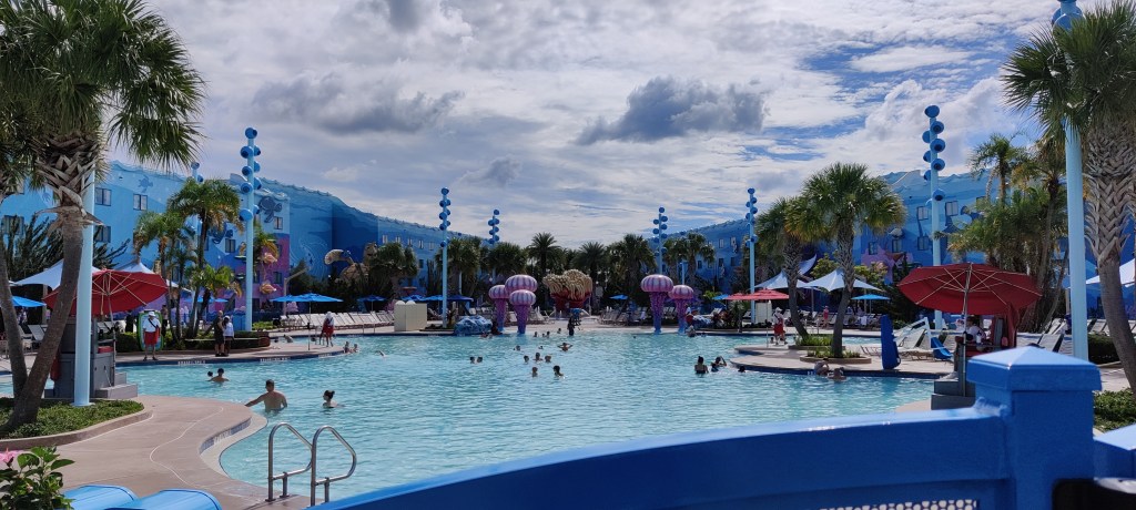 Kuvassa valtava, epämääräisen muotoinen uima-allas, jonka ympärillä on satunnaita päivänvarjoja. Uimassa on jokunen ihminen. Kaukana erottuu medusanmallisia violetteja patsaita. Ilma on pilvinen ja aluetta reunustavat hotellin seinät sinisiä kuten altaan vesi.