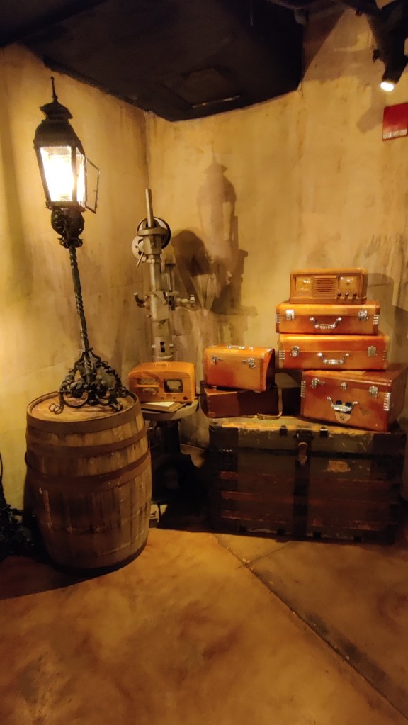 Hämärästi valaistu ravinolan nurkkaus, jossa tynnyrin päällä seisoo matala katulamppu. Sen viereen on pinottu erilaisia ruskeita matka-arkkuja.