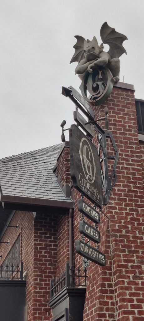 Kuvassa tiilirakennuksen korkea kulma, jossa on puinen kyltti: Gideon's bakehouse, cookies, vakes, curiosities. Rakennuksen kulman, vähän kuin savupiipun päällä, istuu harmaa siivekäs patsas, gargoili, joka näyttää nyrpeää naamaa ja nojailee G-kirjaimeen.