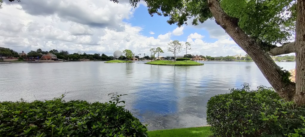Kuvassa vihreiden pensaiden ja puiden reunustama järvialue, jonka keskellä on pari pientä saarta muutamine puineen. Vastarannalla näkyy puita ja rakennuksia sekä iso, valkoinen pallo. Taivas on pilvinen.