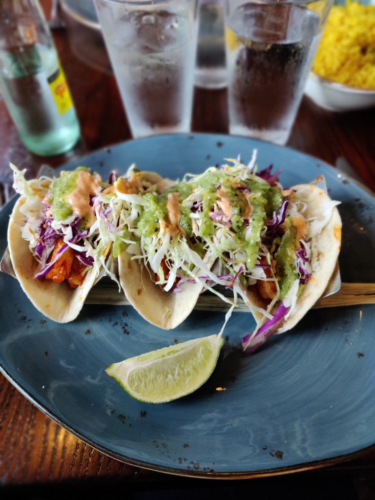 Kuvassa lautasella kolme tacoa, joissa paljon kasviksia ja päällä guacamolea. Vieressä limesiivu.