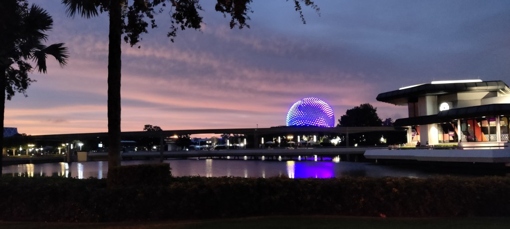 Kuva auringonlaskusta EPCOTissa: keskiessä violetin värinen pyöreä pallo, joka erottuu silhuettimaisen sillan takaa.