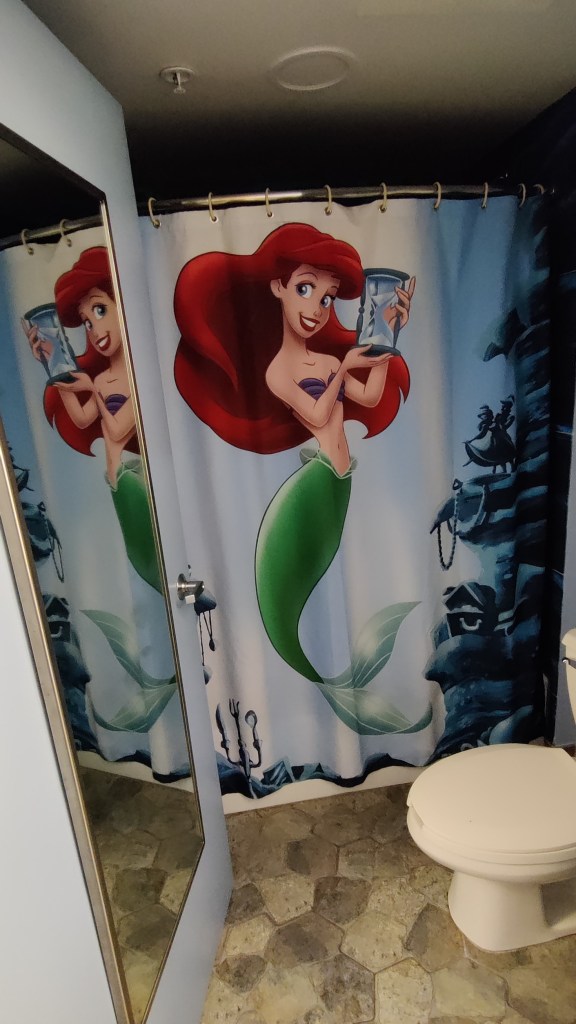 Kuva pienenoloisesta kylppäristä, jonka ovessa on iso kokovartalopeili. Oikealla näkyy valkoinen pönttö, mutta kuvaa hallitsee levitetty suihkuverho, jossa pieni merenneito Ariel pitelee kädessään tiimalasia.