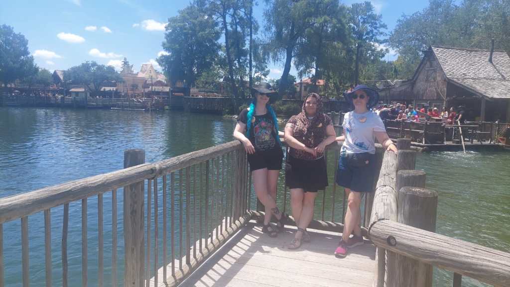 Ryhmäkuvassa Katja, Sari ja Tarja nojailevat puisen laiturin kaiteeseen. Taustalla näkyy lampi ja puisia rakennuksia.