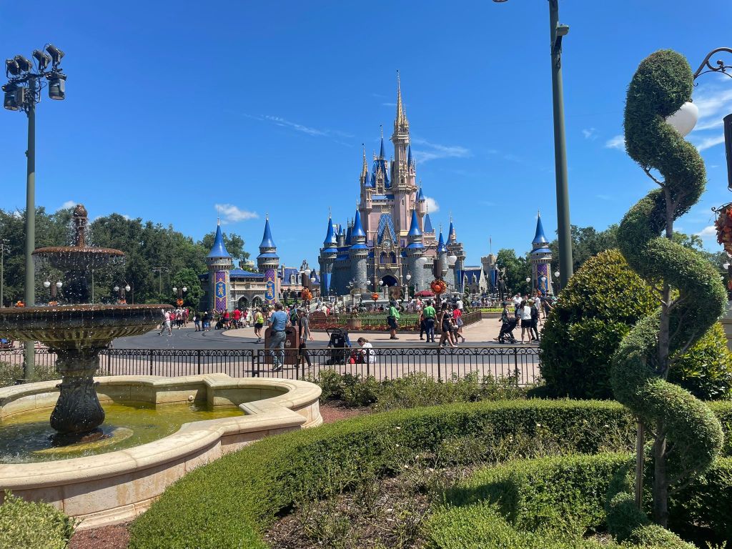 Pienen puistoalueen läpi otettu kuva Tuhkimon linnasta. Puiston pensaat on leikattu tarkkaan muotoon ja suihkulähte pulppuaa vasemmassa reunassa. Ihmisiä on jonkin verran liikkeellä. Linna on vaaleanpunainen ja siinä on useita kapeita tornejä, joiden huiput ovat sinisiä. Taivas on sininen ja lähes pilvetön.