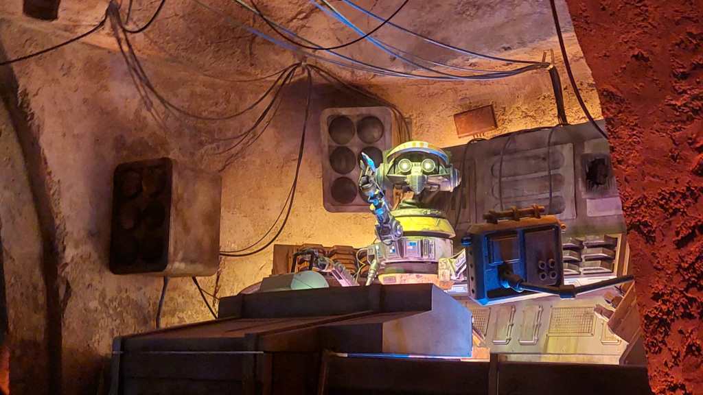Kuvassa luolamaisessa syvennyksessä pöydän takana seisova droidi, joka toimi Oga's Cantinan deejiinä.
