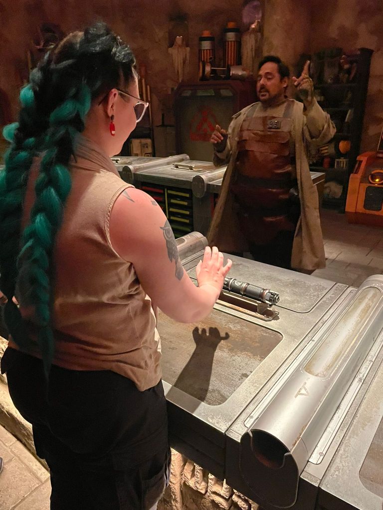 Kuva Katjan selän takaa: Katja pitelee kättään telineelle asetetun kahvan yläpuolella. Pöydän takana on ruskeaan työmiehen esiliinaa pukeutunut tummahiuksinen mies, joka osoittaa kattoa sormellaan.