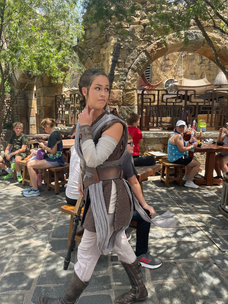 Ihan kameran vierestä kulkent Rey pitkän sauvansa kanssa. Hän katsoo olan yli taakseen kuin varmistaakseen, ettei häntä seurata. Taustalla ihmiset istvat syömässä eväitä puisten pöytien ääressä.