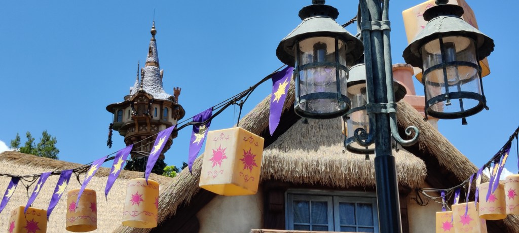 Erikoislähikuvassa katulamppu, josta roikkuu johto, johon on kiinnitetty lukuisia keltaisia lyhtyjä. Lyhdyissä on Tangled-elokuvasta tuttu aurinkologo. Taustalla on olkikatoinen talo, jonka takaa kohoaa Rapunzelin torni.