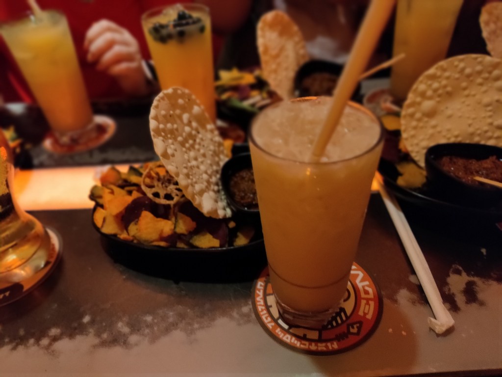 Kuvassa etualalla oranssi juoma, jossa pilli ja jäitä. Sen ympärillä on pari mustaa lautasta täynnä kuivattuja vihanneslastuja. Taustalla näkyy muitakin oransseja juomia, joista yhdessä on mustia helmiä.