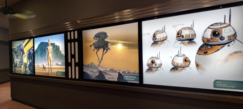 Kuvassa seinällä olevat valaistut ruudut, jossa esillä konseptitaidetta Destroyer-droidista, C-3PO.sta, imperiumin jäljittäjädroidista ja BB-8:sta.