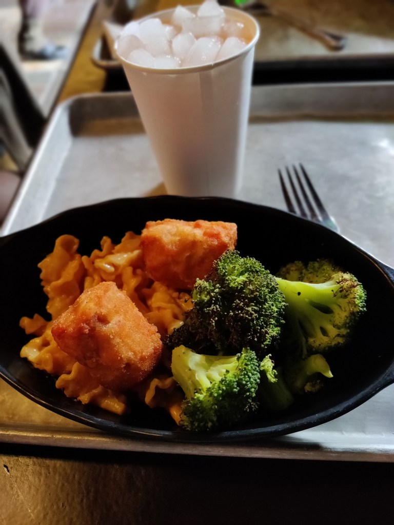 Lähikuva ruoka-annoksesta, jossa mustalla lautasella juustomakaronia, pari isoa friteerattua kananpalaa ja jättimäisiä parsakaaleja. Taustalla muki jäistä juomaa.