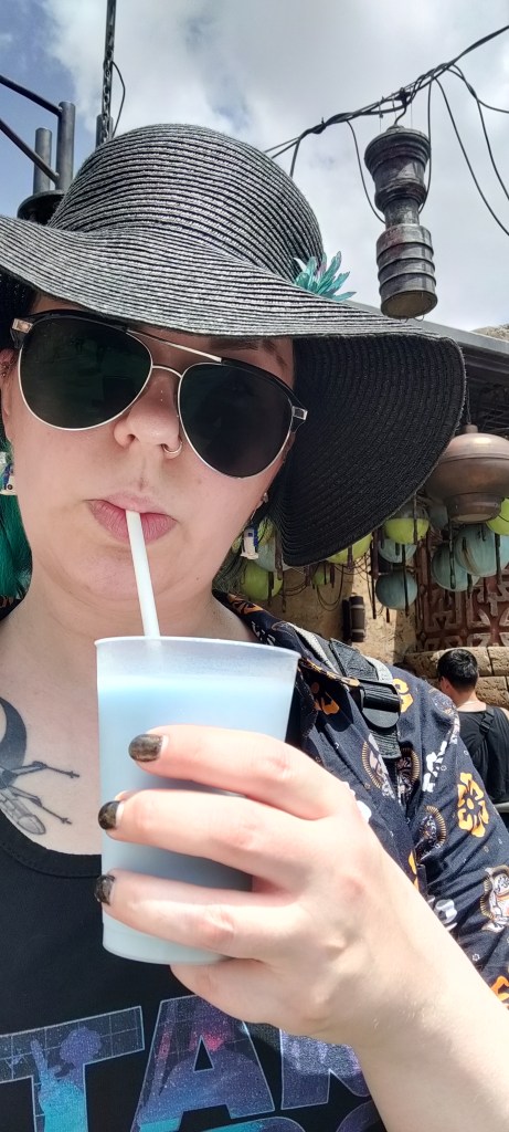 Selfie, jossa Katja juo pillillä muovituopista sinistä maitoa. Katjalla on iso musta lierihattu, pilottihenkiset aurinkolasit ja päällä Star Wars -toppi ja -pusero.