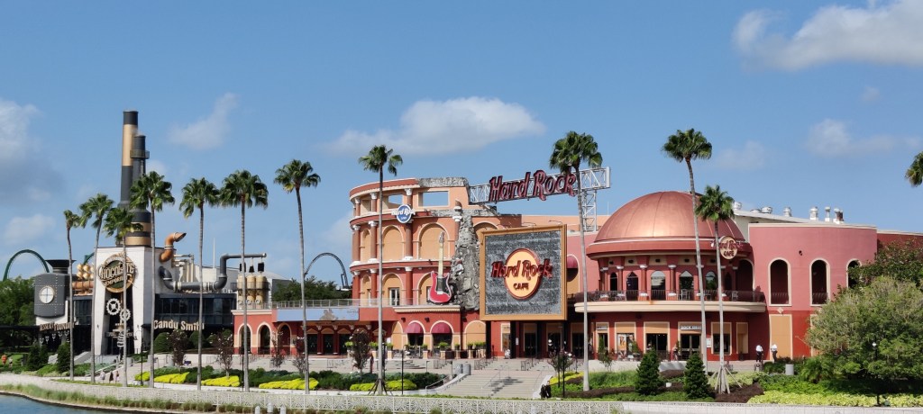 Laaja maisemakuva, jossa vaaleanpunasävyinen iso rakennus pyöreine kupoleineen ja hieman amfiteatterimaisine sivurakennuksineen. Keskellä iso Hard Rock Café -kyltti. Kauempana vasemmalla näkyy hieman epäselvemmin steampunk-henkinen tehdasrakennelma, joka hakee hieman Willy Wonkka -suklaatehdas -tyyliä. Etualalla on muutamia palmuja.