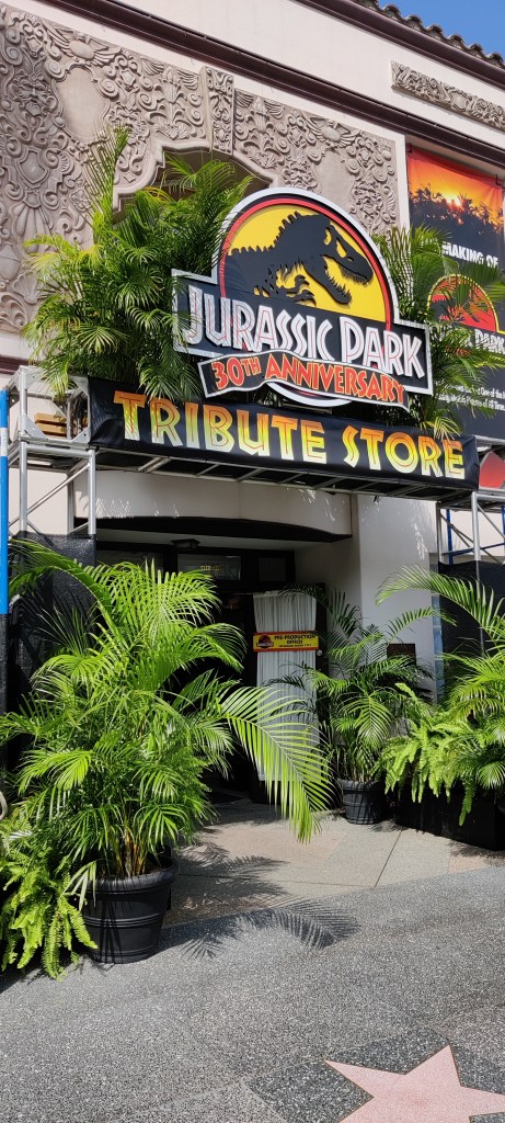 Kuva Jurassiv Park 30th Anniversary Tribute Storen ulkosivusta, jota hallitsee iso määrä viherkasveja ja suuri Jurassic Parkin logo.