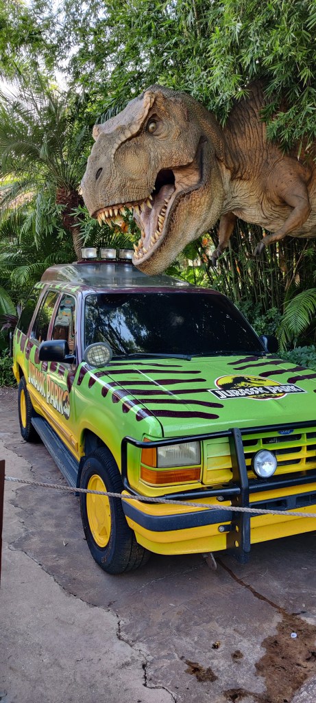 Kuvassa ensimmäisestä Jurassic Park -elokuvasta tuttu keltaisenvihreä Jeeppi, jonka ylle on kumartuneena pusikosta puoliksi näkösällä oleva Tyrannosaurus Rex.