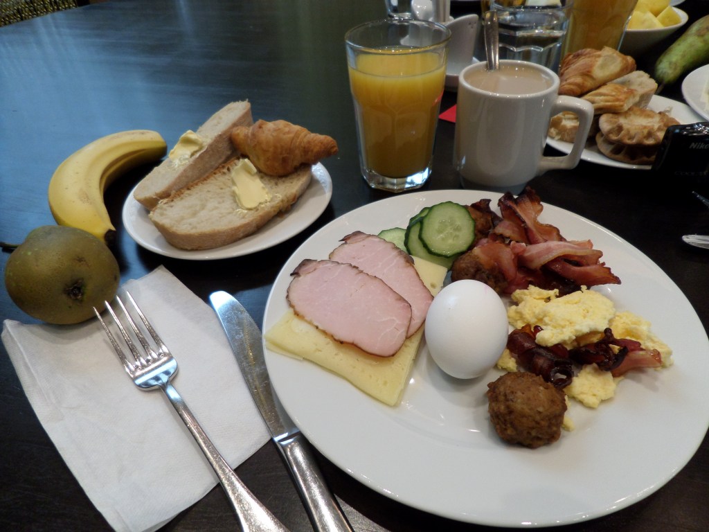 Kuvassa ruokalautanen täynnä pekonia, munakasta, lihapullia, kananmunaa, kinkku- ja juustosiivuja sekä kurkkua. Lautasen vieressä ovat ruokailuvälineet, servetti, päärynä ja banaani. Pienellä lautasella taaempana on kaksi vajaasti voideltua leipäsiivua ja croissantti. Lasissa on appelsiinimehua ja mukissa teetä.