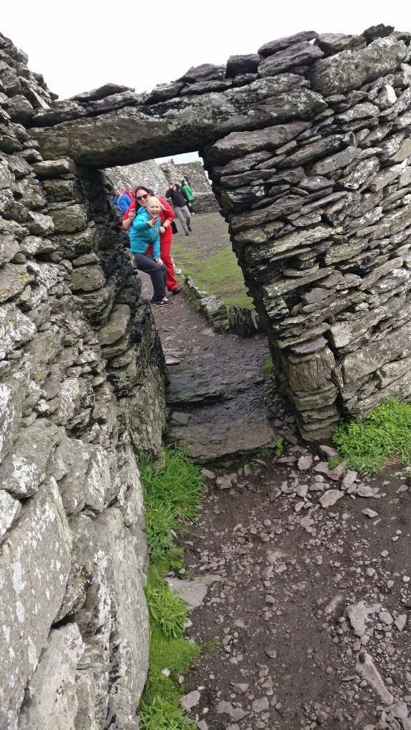 Kuva litteista kivistä kasatusta muurista, jossa on suorakulmainen oviaukko. Oviaukon läpi näkyy kaksi ystävääni, jotka nauravat leveästi ja näyttävät sormillaan rauhanmerkkejä kallistuneena kumaraan. Oviaukko on vino, joten kaikki näyttää olevan vinossa.