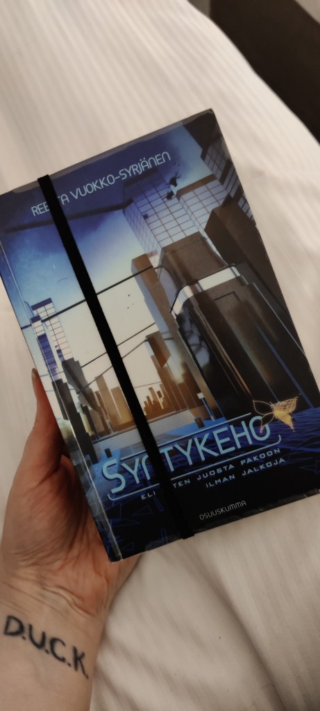 Katja pitelee kädessään pokkaria, jonka kannen edestä kulkee pystyssä musta kuminauha.