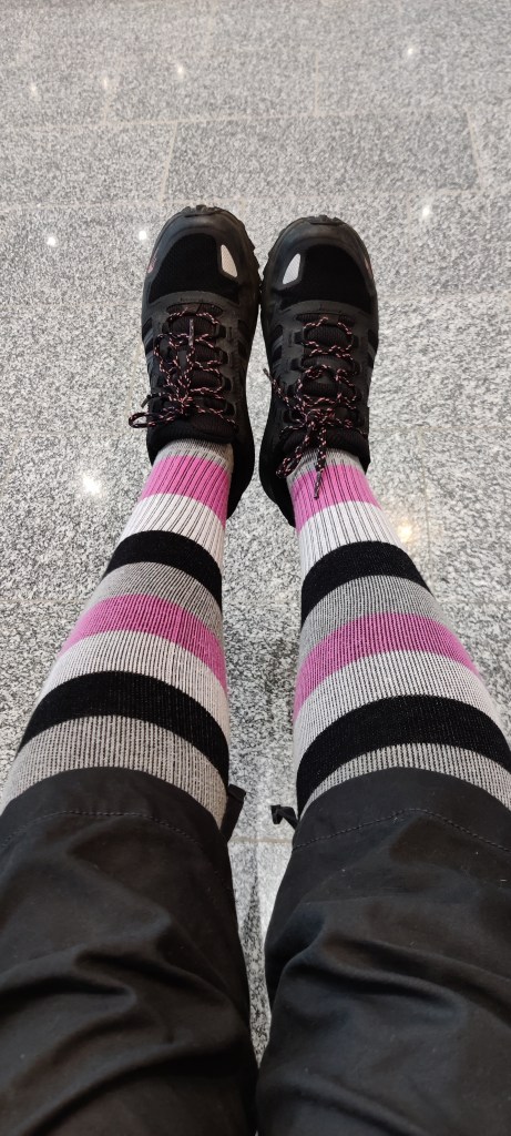 Kuvassa Katjan jalat polvista alaspäin. Polvien alle yltävien löysien caprien alta paljastuvat valkomustapinkkiraidalliset tukisukat. Jalassa on mustat vaelluskengät.