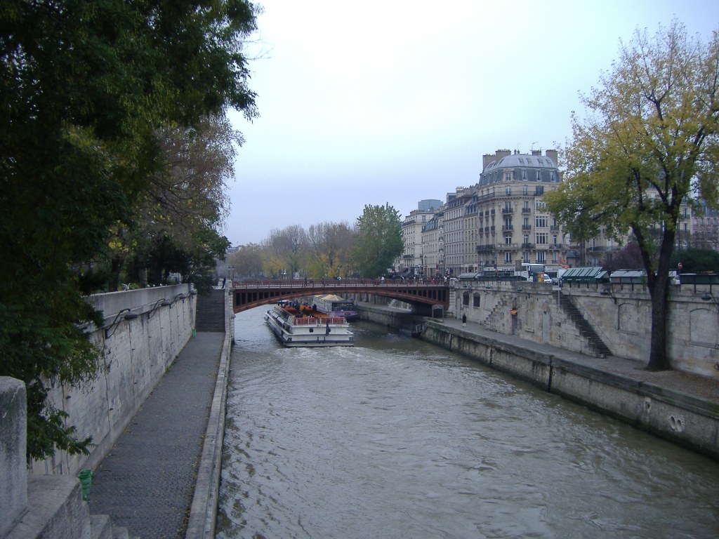 Kuvassa kapea, rakennettujen kävelykatujen keskellä kulkeva joki, jolla matala jokilaiva seilaa poispäin kamerasta. Laiva on juuri matalan sillan alla. Vasemmassa reunassa on paljon puita, kuten myös edessä korisontissa. Oikealla seisoo yksi puu, jonka lehdet ovat alkaneet vaalentua. Taustalla näkyy korkeita, vanhoja rakennuksia.