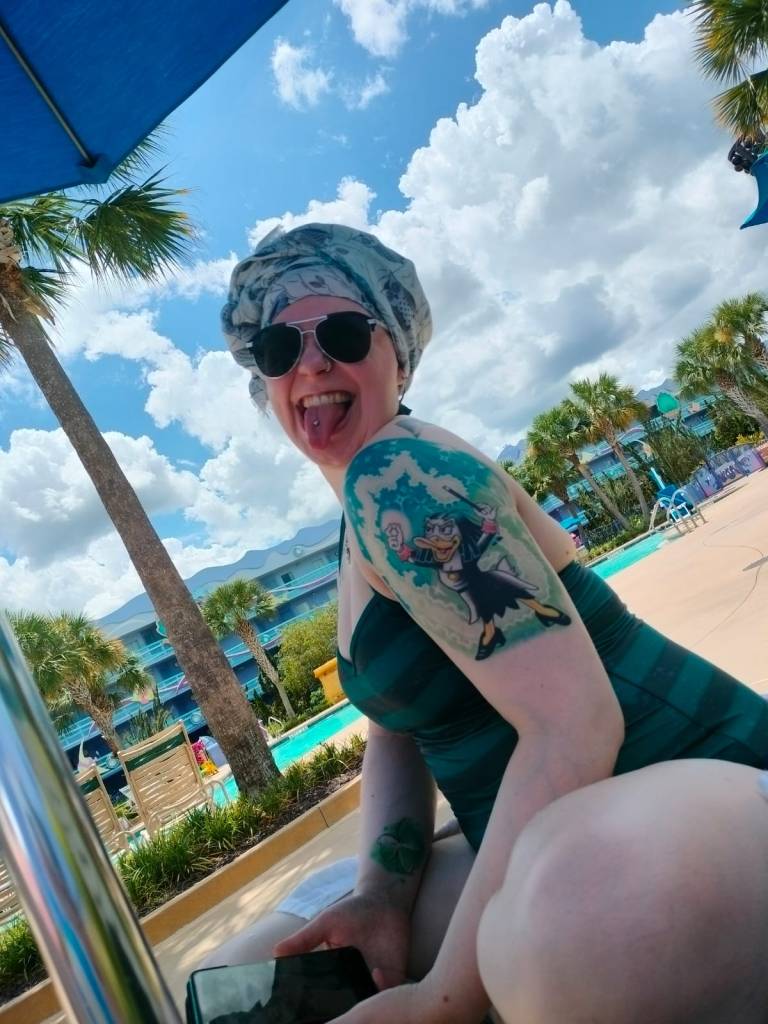 Kuva Katjasta, joka näyttää kieltä kameralle. Katjalla on huivi ja aurinkolasit päässä ja hän istuu uimapuvussa uima-altaan lähellä olevalla tuolilla. Kuvassa näkyy aurinkovarjon kulma ja useita palmuja. Kuvan keskiössä on kuitenkin olkapään näyttävä Milla Magia -tatuointi.