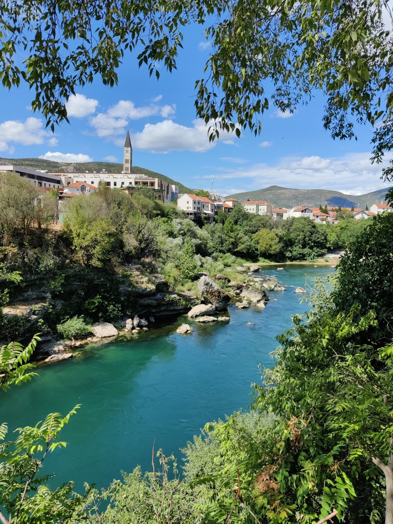 Kuvassa etualalla pensaikkoa ja puiden lehtiä, jotka rajaavat kuvaa alhaalta nousten yläspäin oikeaa reunaa pitkin. Kuvan yläreunasta roikkuu lisää vihreitä puunlehtiä.
Sinivihreänä hehkuva joki kulkee vinottain kuvan poikki alhaalta vasemmalta oikealle. Joen toisella rannalla on kivikkoa ja pensaikkoa, joiden takana näkyy valkeita rakennuksia, joilla on oranssit katot. Yksittäinen kirkontorni kohoaa korkeimmalle. Sää on aurinkoinen, hieman pilvinen. Taustalla kumpuilevaa maastoa.