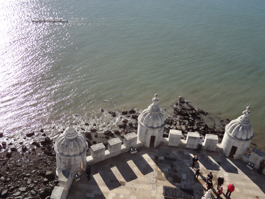 Ylhäältä alaspäin otettu kuva, jossa alareunassa on pyöreänmallisen terassin reunat mataline tornikoristeineen. Terassin ympäriltä näkyy rantakivikkoa ja valtaosa kuvasta on harmaatä vettä, joka kimaltelee auringossa kuvan vasemmassa reunassa. Terassilla näkyy muutamia ihmisiä ja vasemmassa ylänurkassa menee soutuvene, jossa on useita soutajia.