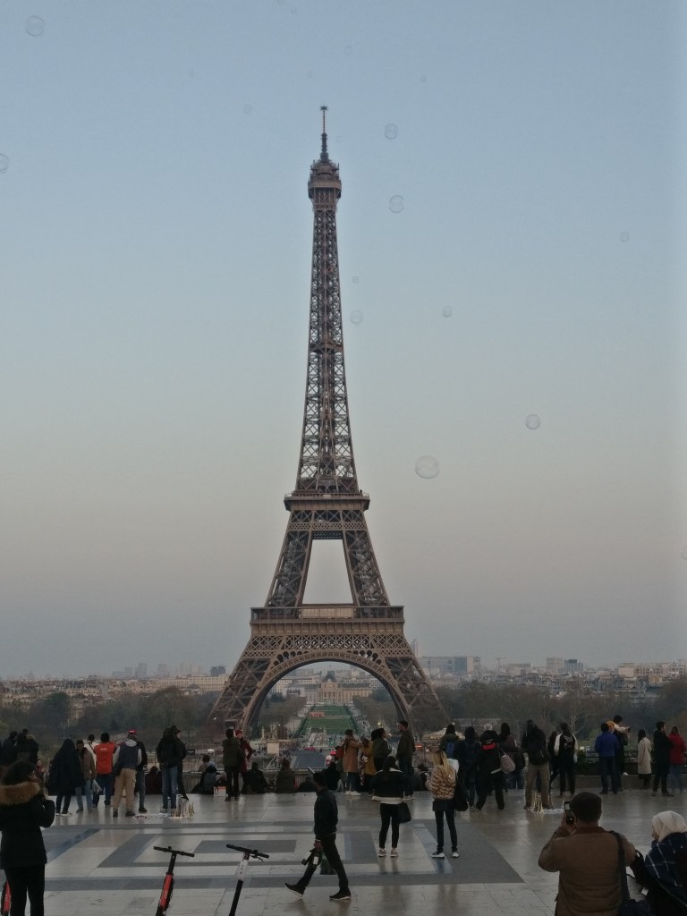 Kuvassa keskellä massiivinen Eiffel-torni, jota lukuisa määrä ihmisiä kuvaa kameroillaan. Kuvassa on vesitäpliä, jotka ovat jääneet kameran linssistä koristamaan harmaata taivasta.