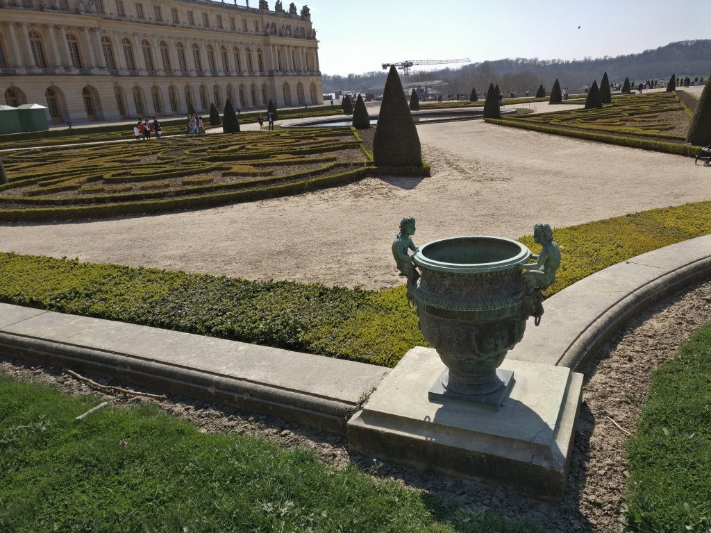 Kuvassa etualalla sinieksi tummunut maljakko, jonka reunoista pitelee kaksi pientä ihmishahmoa. Maljakko on matalan aitauksen kulmassa ja sen takaa levittyy geometrinen puutarha hiekoitettuine kävelyalueineen. Pensasaidat ovat vielä aivan matalia. Taustalla näkyy Versailles'n palatsin päätyä. Kauempana kävelee joitain ihmisiä.