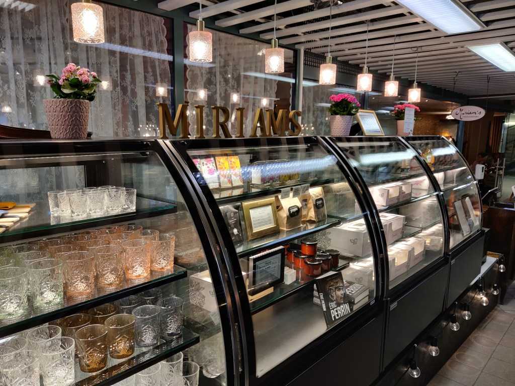 Kuvassa Miriam'sin myyntitiskit käytävällä kahvilan ulkopuolella. Tiskissä on myynnissä koristeellisia juomalaseja, paikallisten pienryrittäjien tuotteita, kuten Papu-paahtimon kahvia, sekä oman konditorian kakkuja.