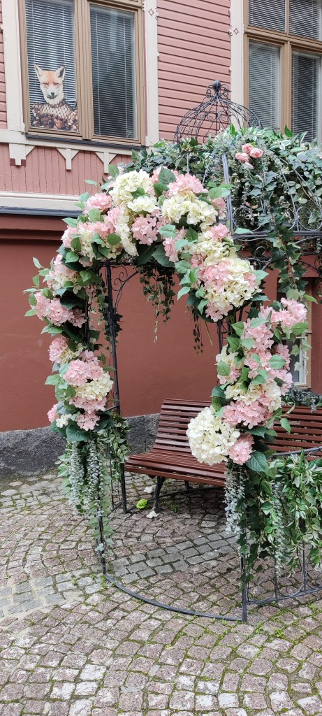 Kuvassa Nikolainkulman sisäpihalla oleva rautainen, iso "lintuhäkki", jonka sisällä on ruskea puistonpenkki. Lintuhäkki on koristeltu isoilla kukkaistutuksilla ja näyttää satupuutarhasta karanneelta.