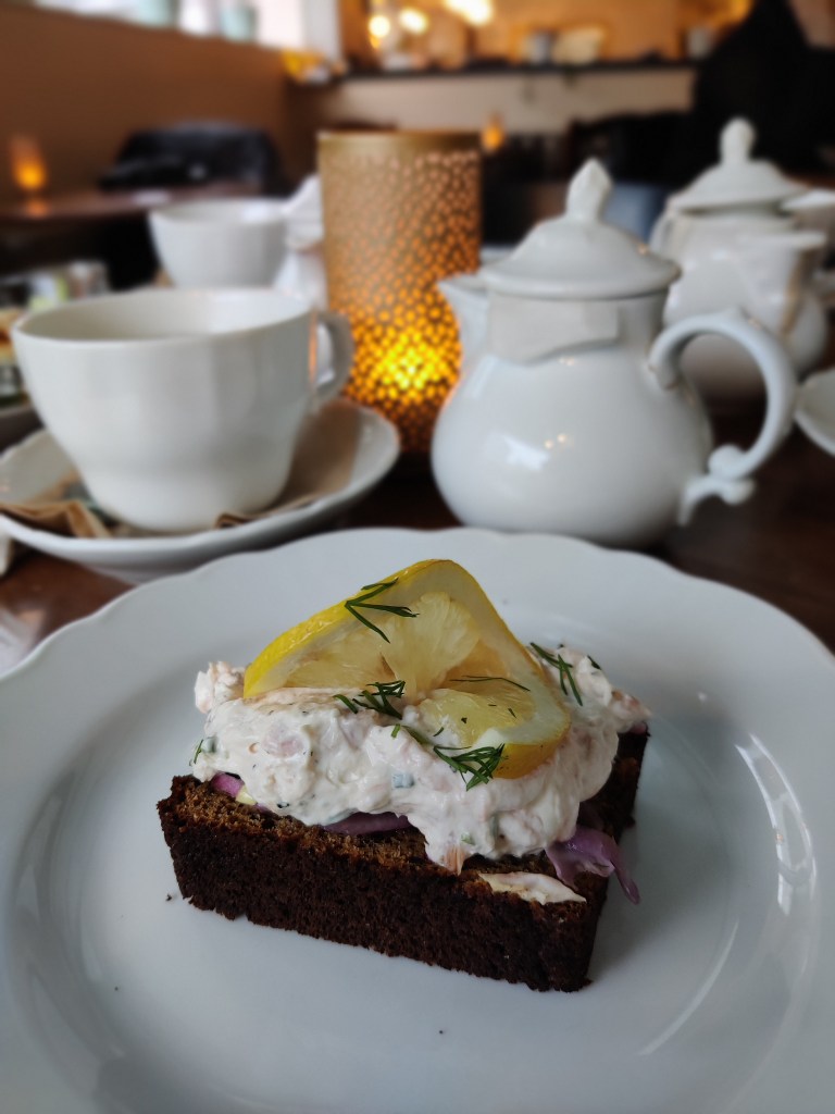 Kuvassa etualalla saaristolaisleipä, jonka päällä on lohimoussetäyte ja sitruunansiivu. Pöydällä on myös pieniä valkoisia teepannuja sekä teekuppeja asetteineen.