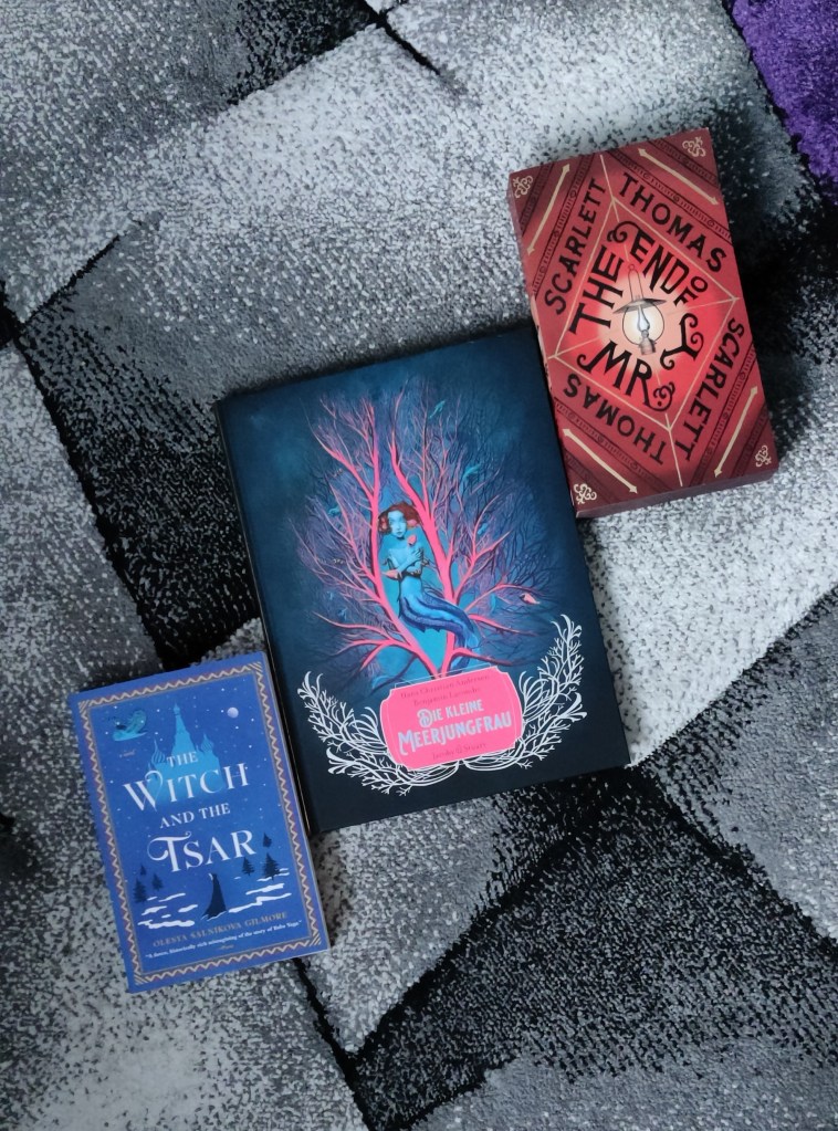 Kuvassa matolle levitettynä kolme kirjaa: The Witch and the Tsar -romaani, Die Kleine Meerjungfrau -kuvakirja, ja The End of Mr. Y -romaani.