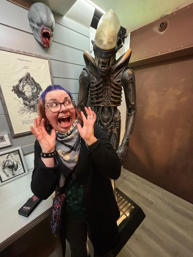 Kuvassa Katja, joka kiljuu mukamas pelokkaana kädet pystyssä ja katsoo sivusilmällä takanaan irvistävää alienia.