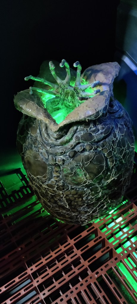 Ritilälattialla seisoo luonnollisen kokoinen alienin muna, josta on juuri kuoriutumassa facehugger. Kelmeä vihreä väri korostaa uhkaavaa tunnelmaa.
