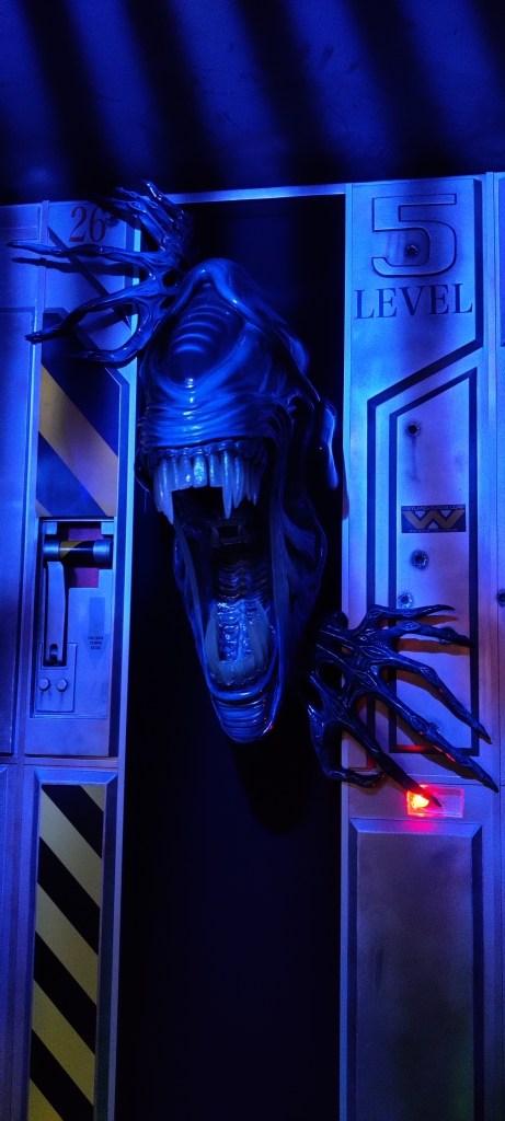 Kuvassa hämärässä valaistuksessa oleva oviaukko, jonka läpi näyttää työntyvän kuningatar-alienin irvistävä pää ja kädet.