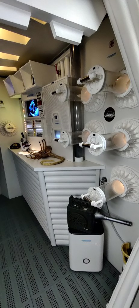 Ashin laboratoriossa seinällä on lukuisia näyteastioita. Pöydällä näkyy selällään retkottava facehugger, mikroskooppi ja lippis. Työpöydän ylöpuolella on tietokoneen näyttö, jossa näkyy jonkinlainen röntgenkuva. Etualalla on Alien-elokuvan liiketunnistin.